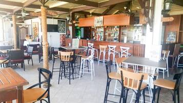 An outdoor cafe with wooden tables and chairs, a bar area, and a tropical palm tree.