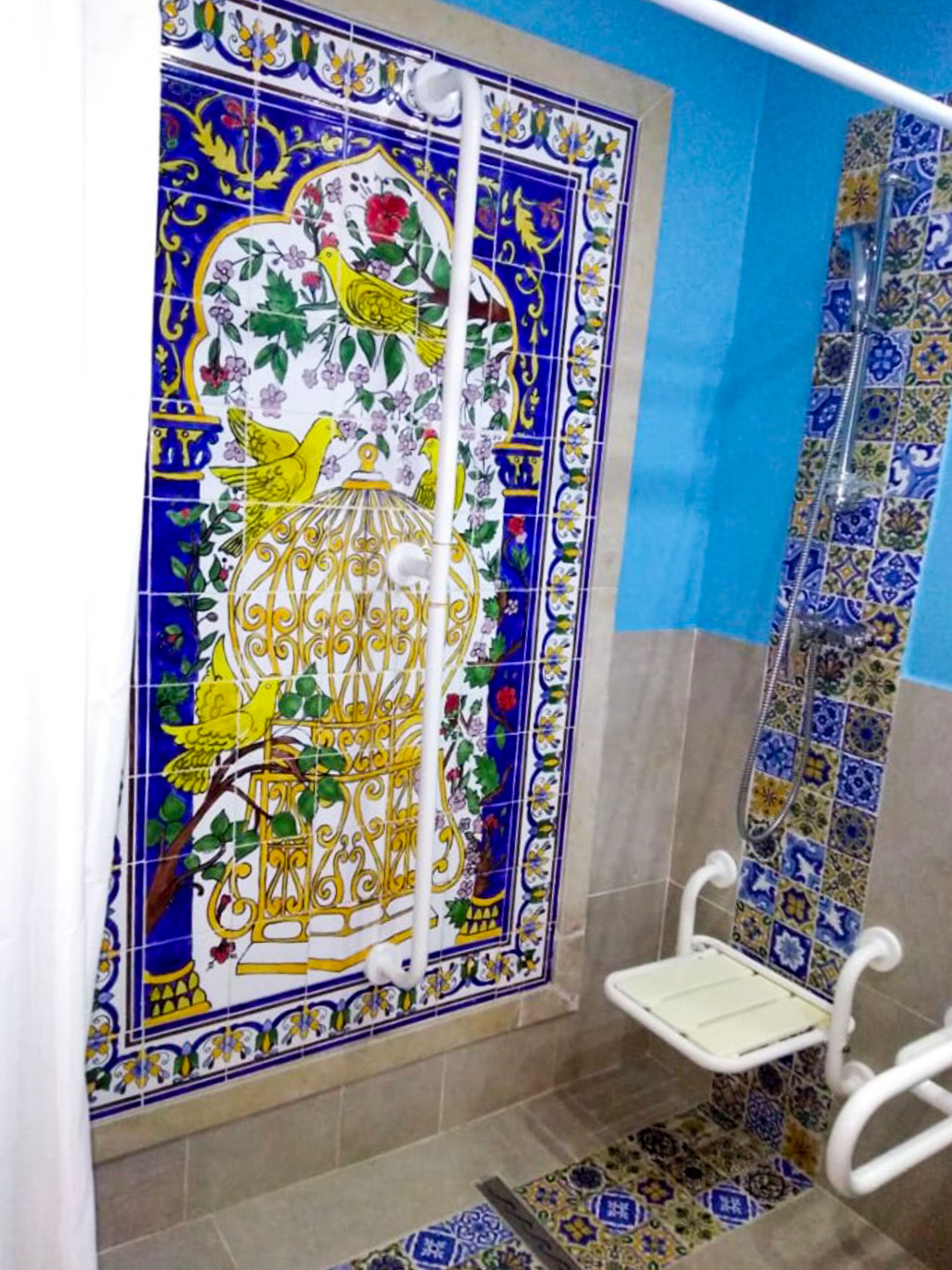 Een badkamer met een douchegedeelte, afgewerkt met blauw-witte betegeling, een witte douchestoel en een decoratieve tegelwand met bloem- en vogelmotieven.