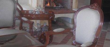 A room with a fireplace, two upholstered chairs, a glass-top table, and a tiled floor.