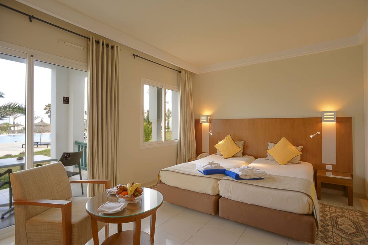 A hotel room with a bed, a chair, a table with fruits, and a view of the outdoors.