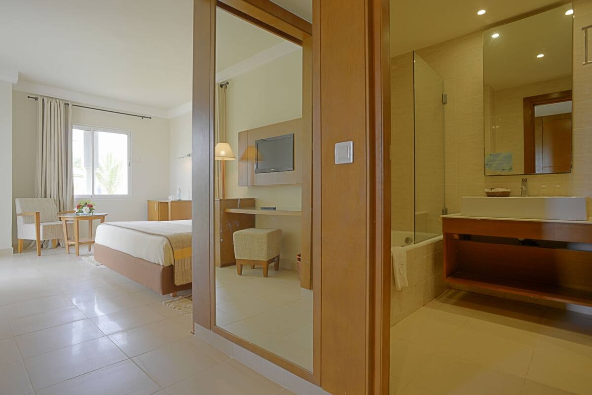 A hotel room with a bed, a desk, a chair, a TV, a bathroom with a mirror, and a window with curtains.