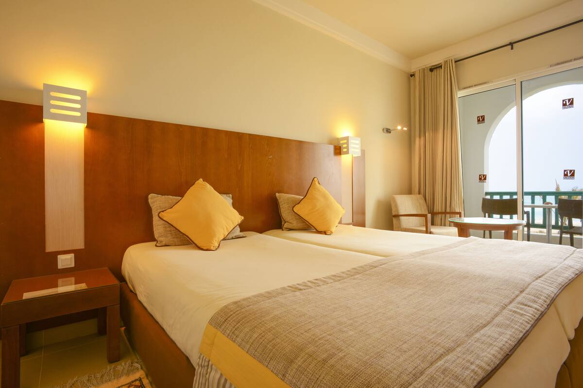 A hotel room with two beds, a nightstand, a chair, a table, and a view of the sea through a large window.