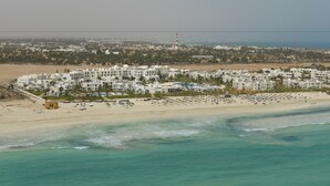 An aerial view of a coastal resort with multiple white buildings, a sandy beach, and a clear view of the sea. - Vincci Helios Beach (Aghir)