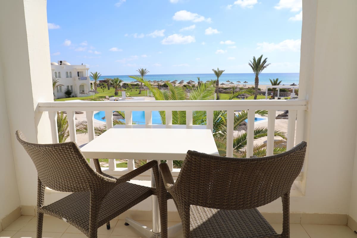 A balcony with wicker chairs and a white table, overlooking a beach and a pool.