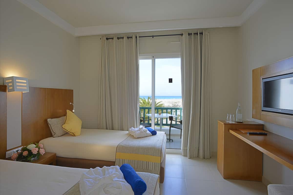 A hotel room with two beds, a TV, a desk, and a balcony with a view of the beach.