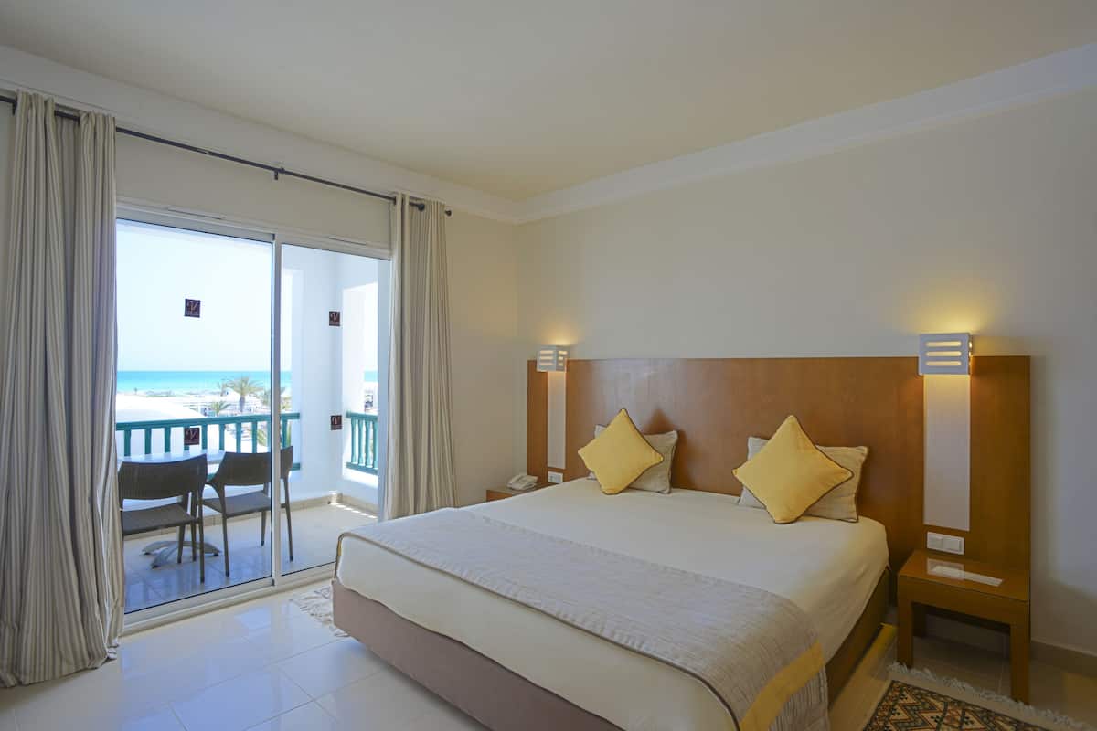 A hotel room with a large bed, two bedside tables, a nightstand, a balcony with a view of the beach, and a chair.