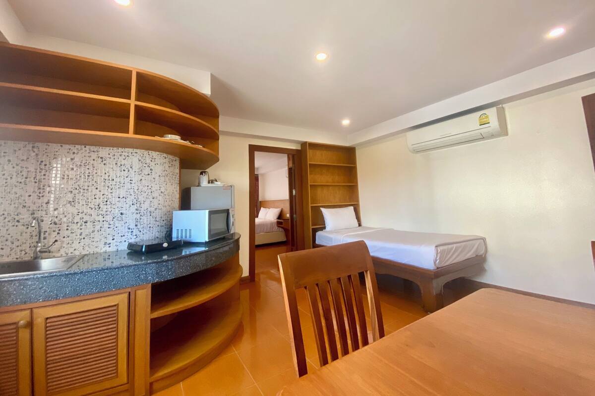 A compact hotel room with a kitchenette, a bed, a wooden chair, and an air conditioning unit.