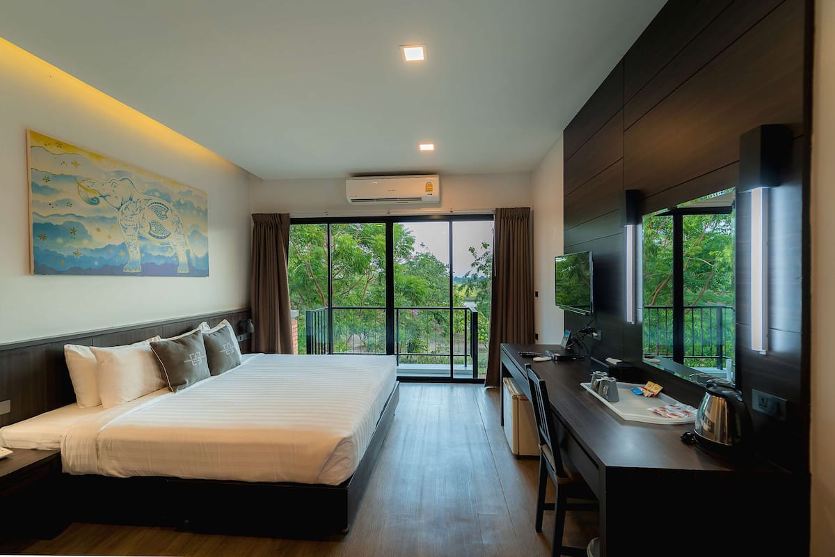 A modern hotel room with a large bed, a desk with a kettle, and a TV. There is a balcony with a view of trees.