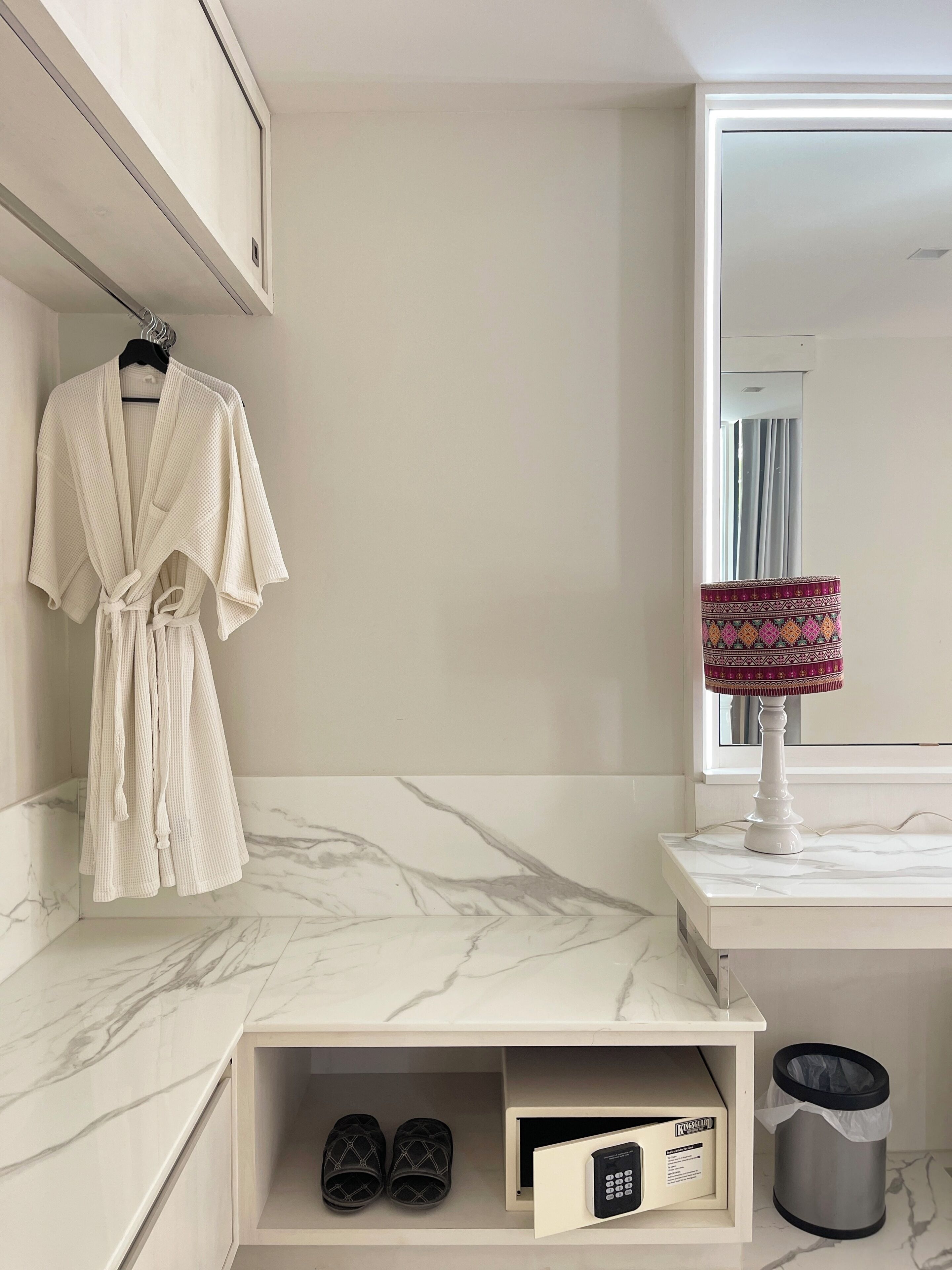 A bathroom with a marble countertop, a robe hanging on the wall, a lamp, a trash can, and a shoe storage unit.
