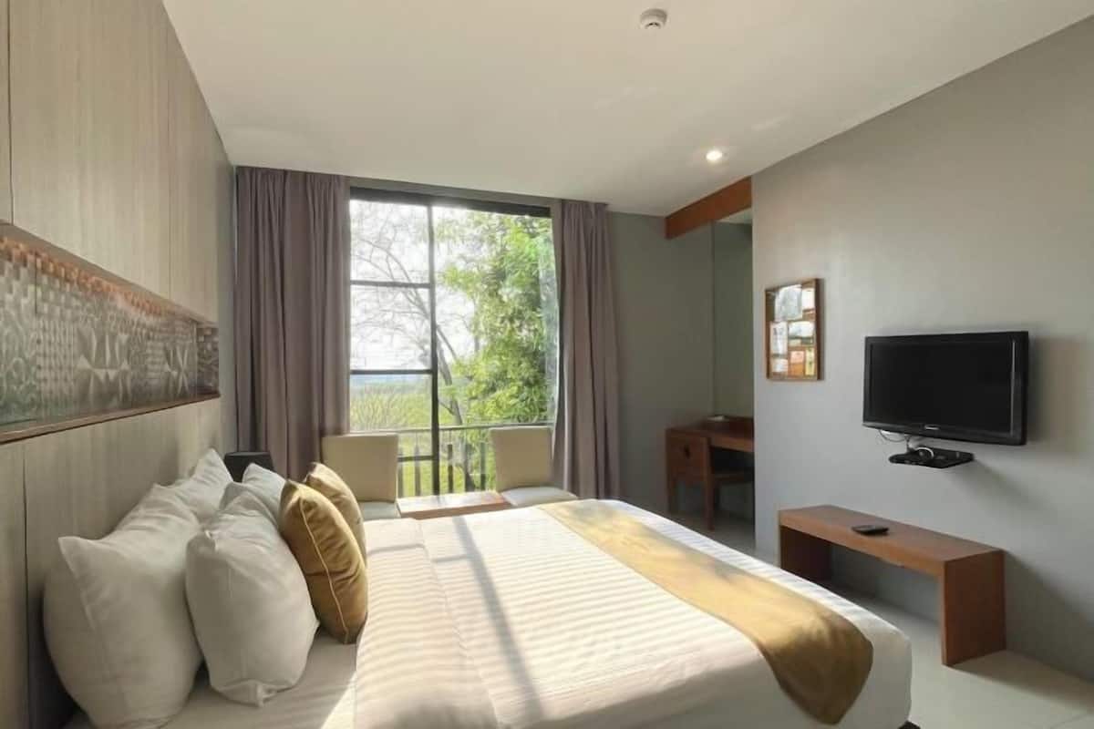 A modern hotel room with a large bed, a desk, a television, and a balcony with a view of greenery.