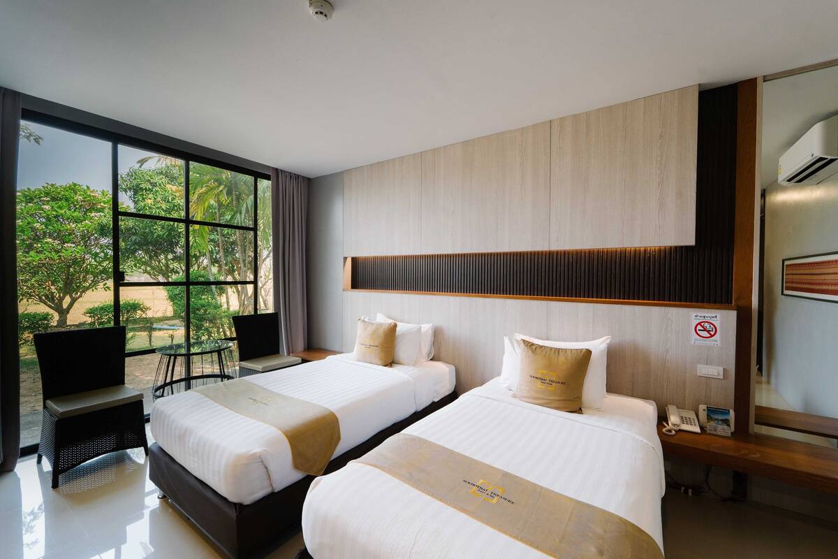 A hotel room with two beds, a large window with a view of greenery, a desk with a phone, and a mirror.