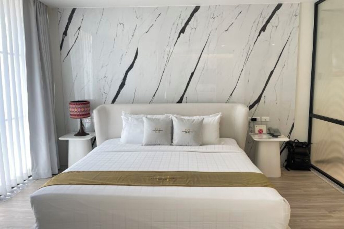 A hotel room with a large bed, a nightstand with a lamp, a small table with a phone, and a view of the outside.