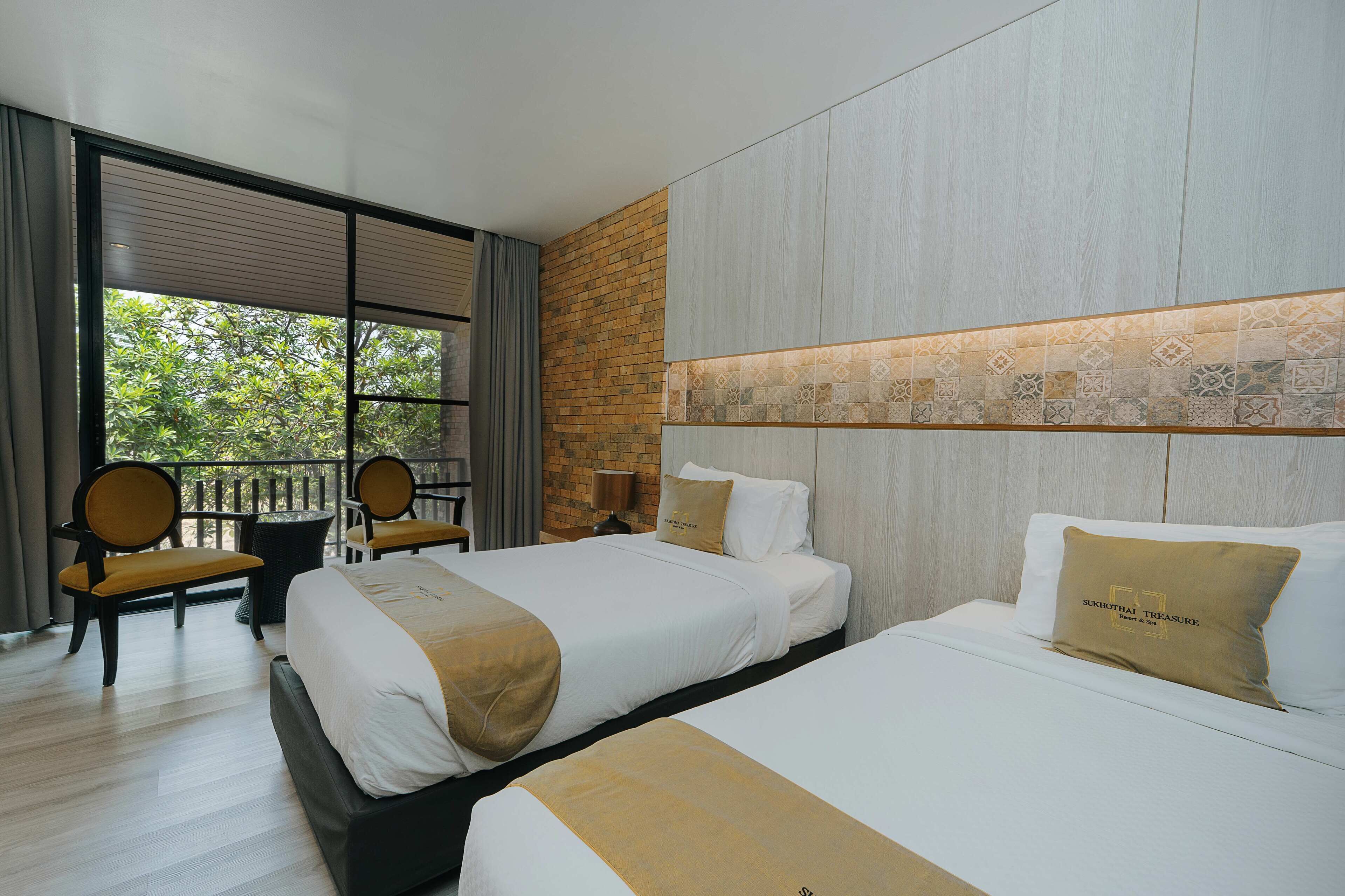 A hotel room with two beds, a balcony with chairs, and a view of greenery.