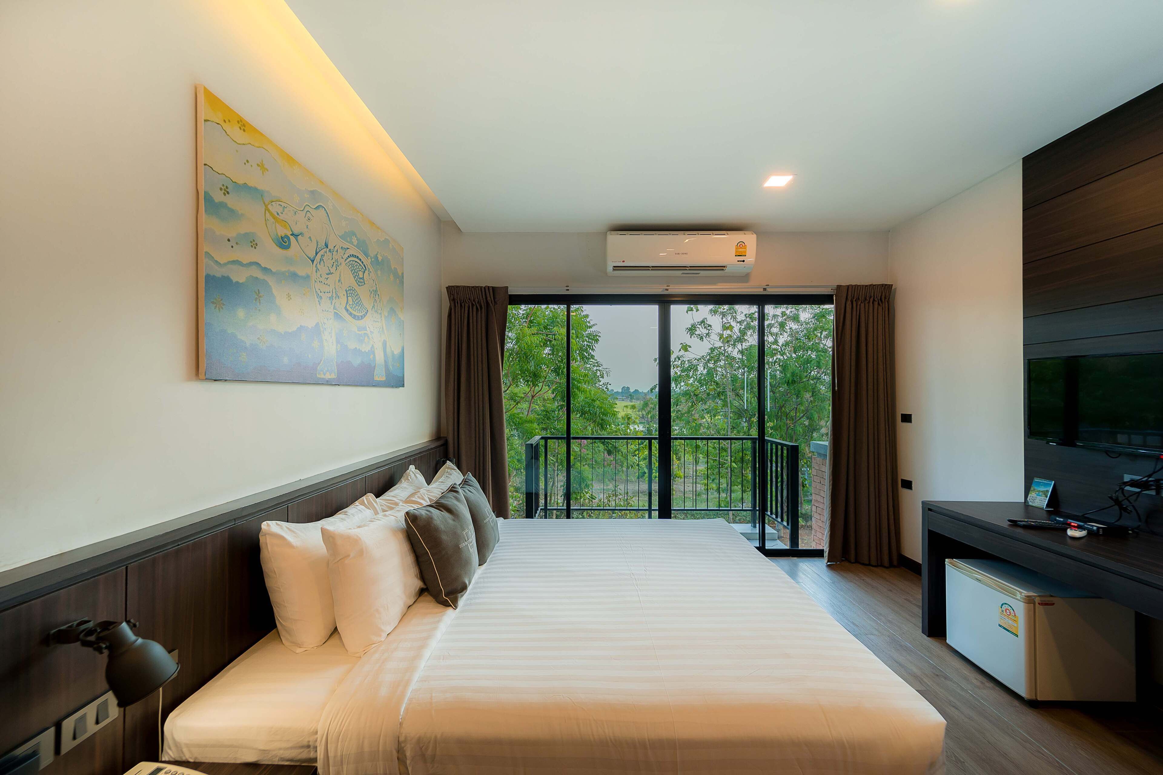 A hotel room with a large bed, a desk, a TV, and a balcony with a view of greenery.
