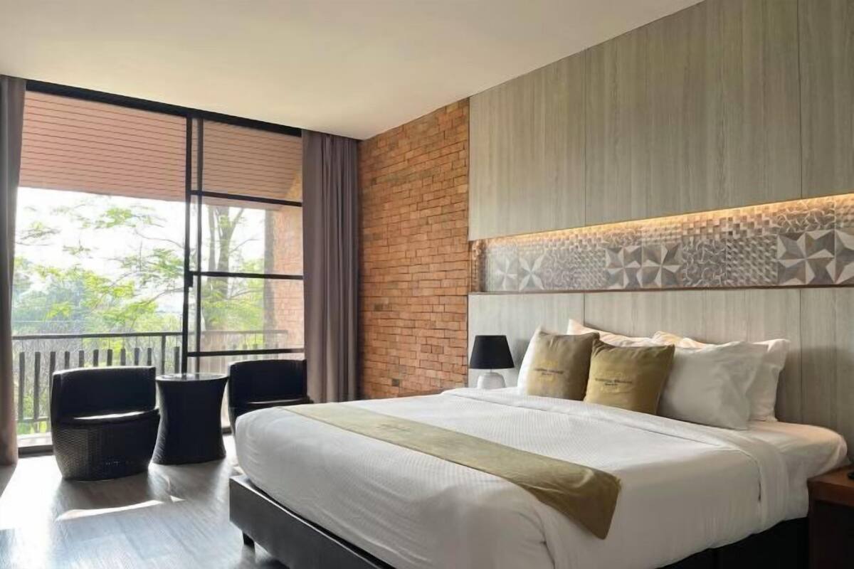 A hotel room with a large bed, a view through the window, a brick wall, and a patterned headboard.