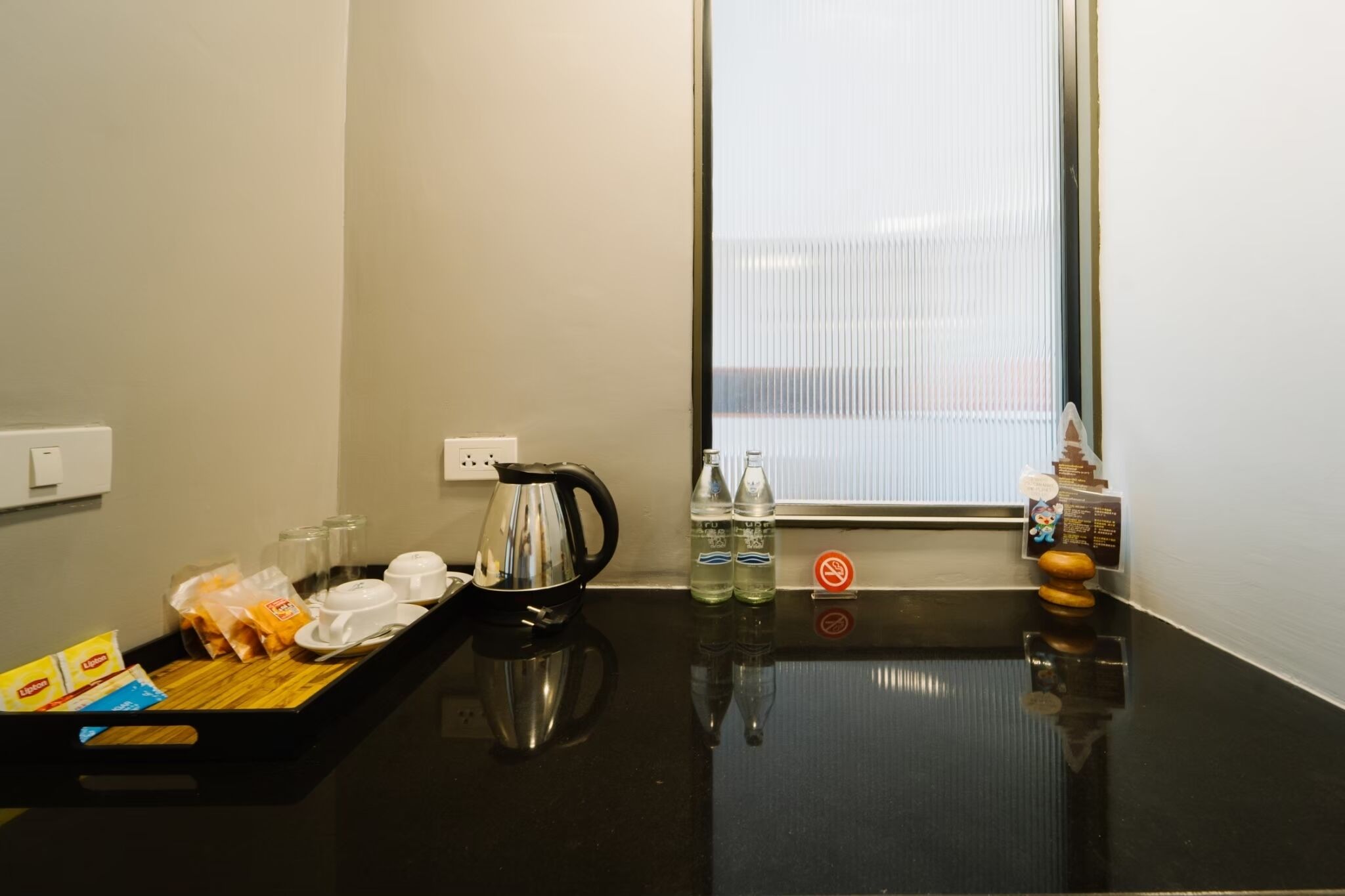 Habitación de hotel con mostrador negro, hervidor de agua, productos para el desayuno y una ventana con persianas.