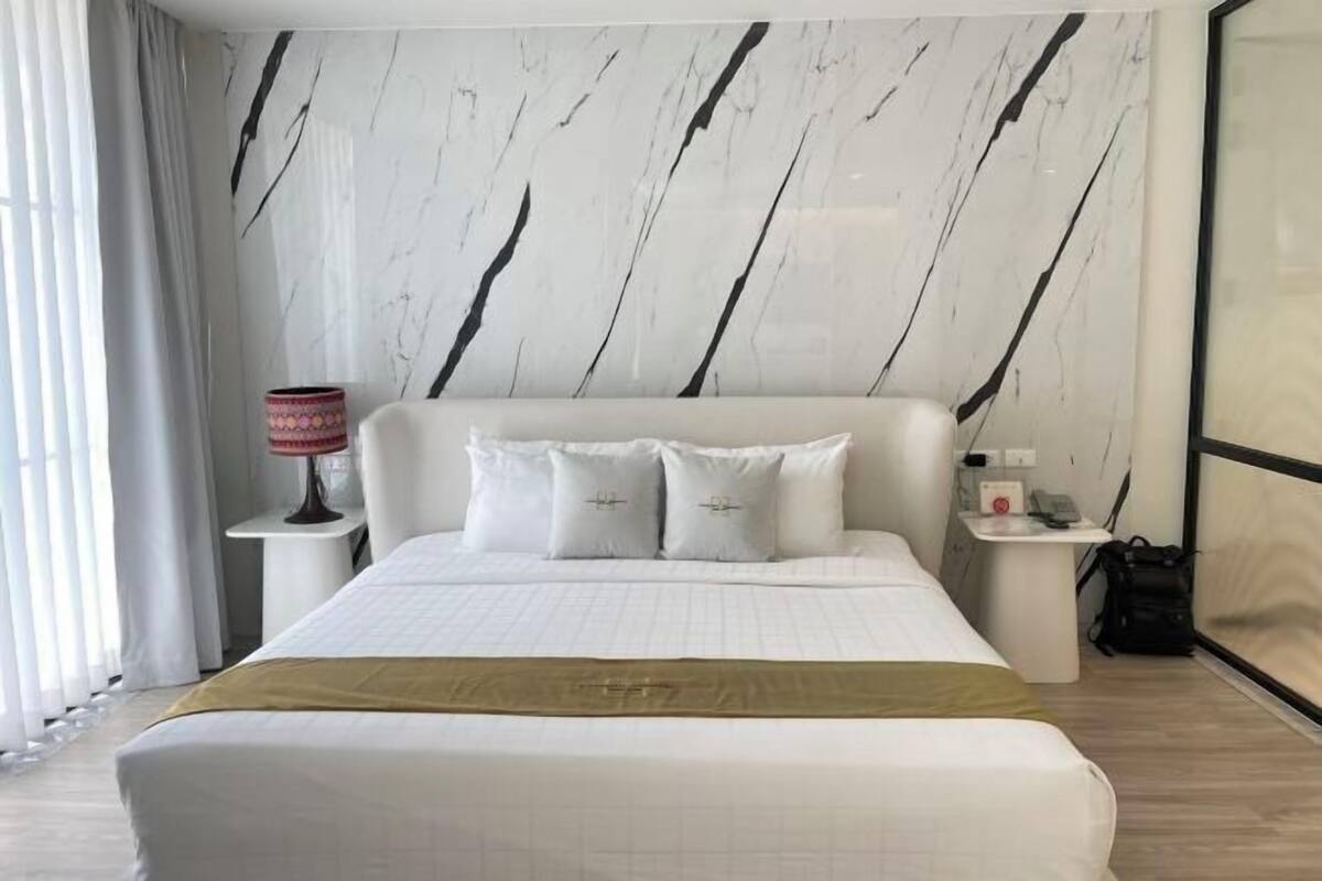 A hotel room with a large bed, a nightstand with a lamp, a small table with a phone, and a view of the outside.