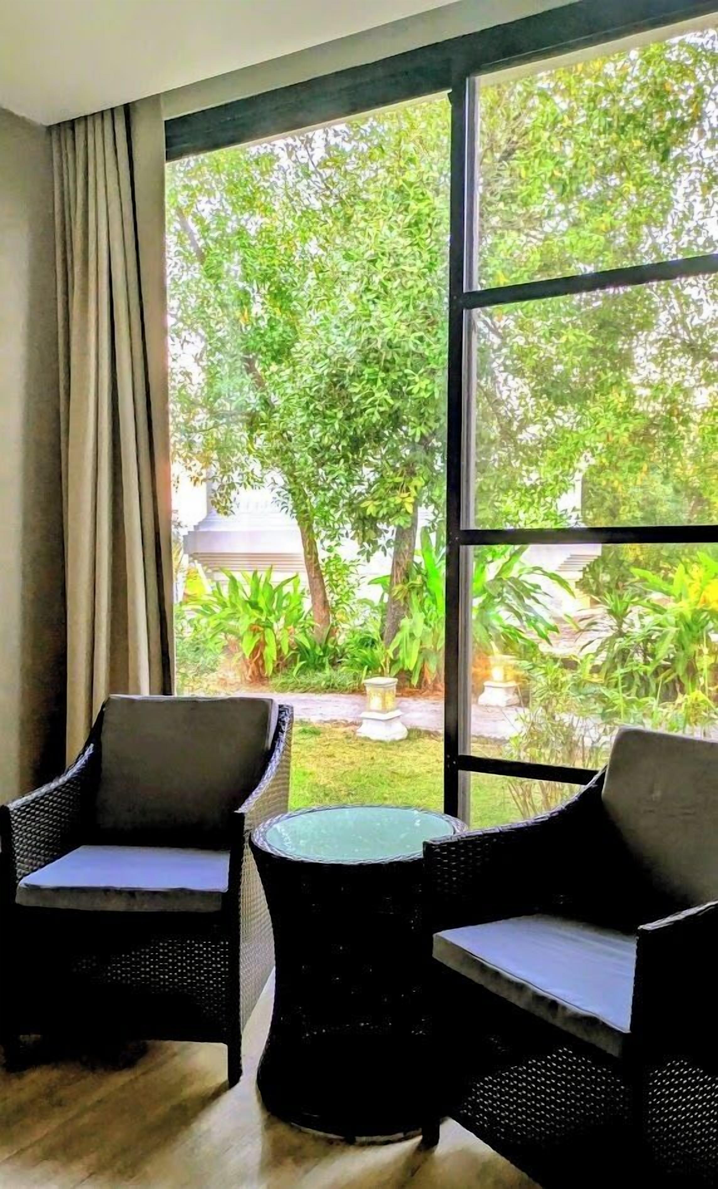 A room with two wicker chairs and a small table, large windows with a view of greenery outside.
