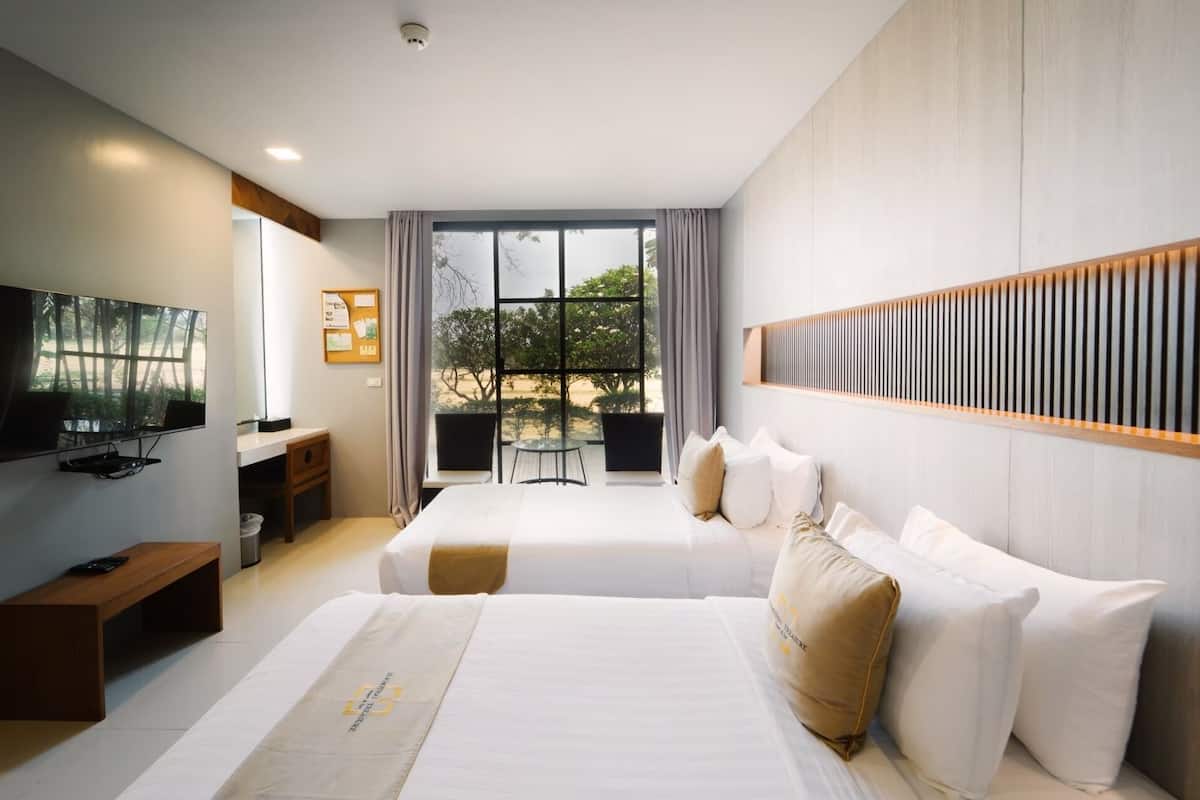 A hotel room with two beds, a TV, a desk, and a balcony with a view of trees and a building.