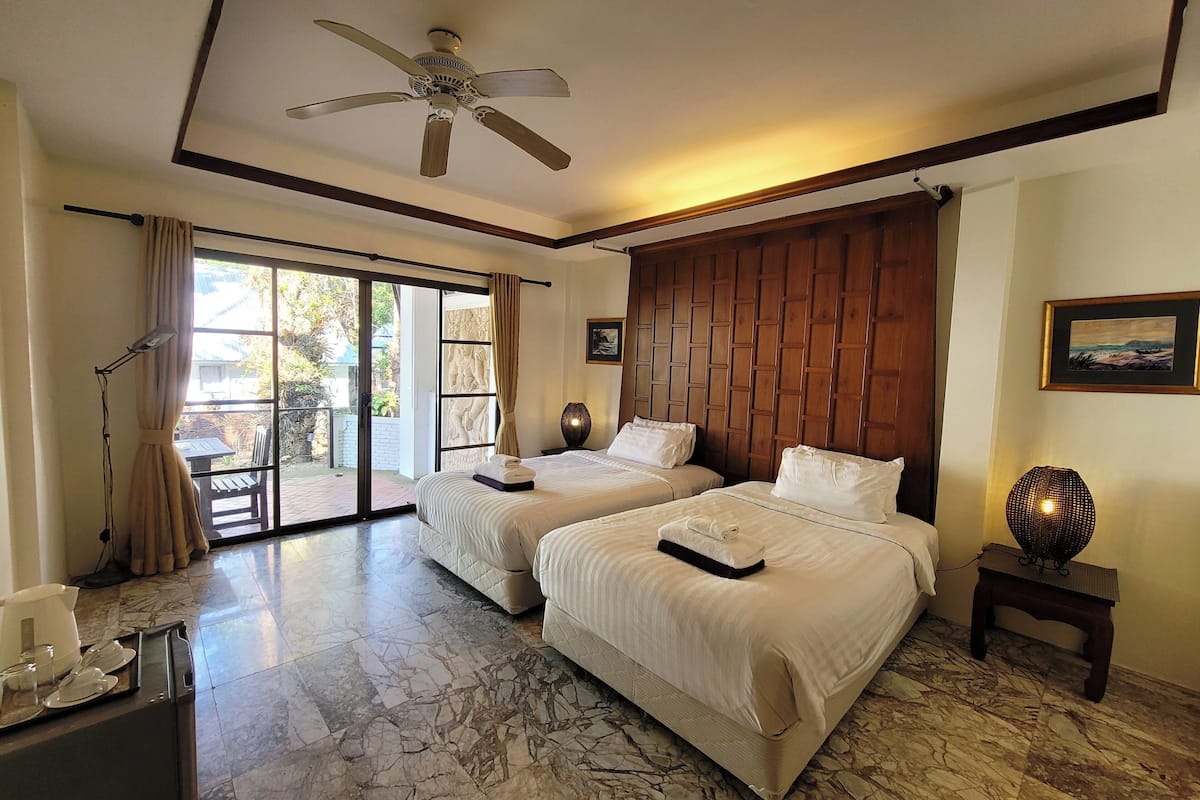 Une chambre d’hôtel avec deux lits, un ventilateur de plafond, un coin repas avec une table et des chaises, et une vue sur l’extérieur grâce à une porte-fenêtre coulissante.