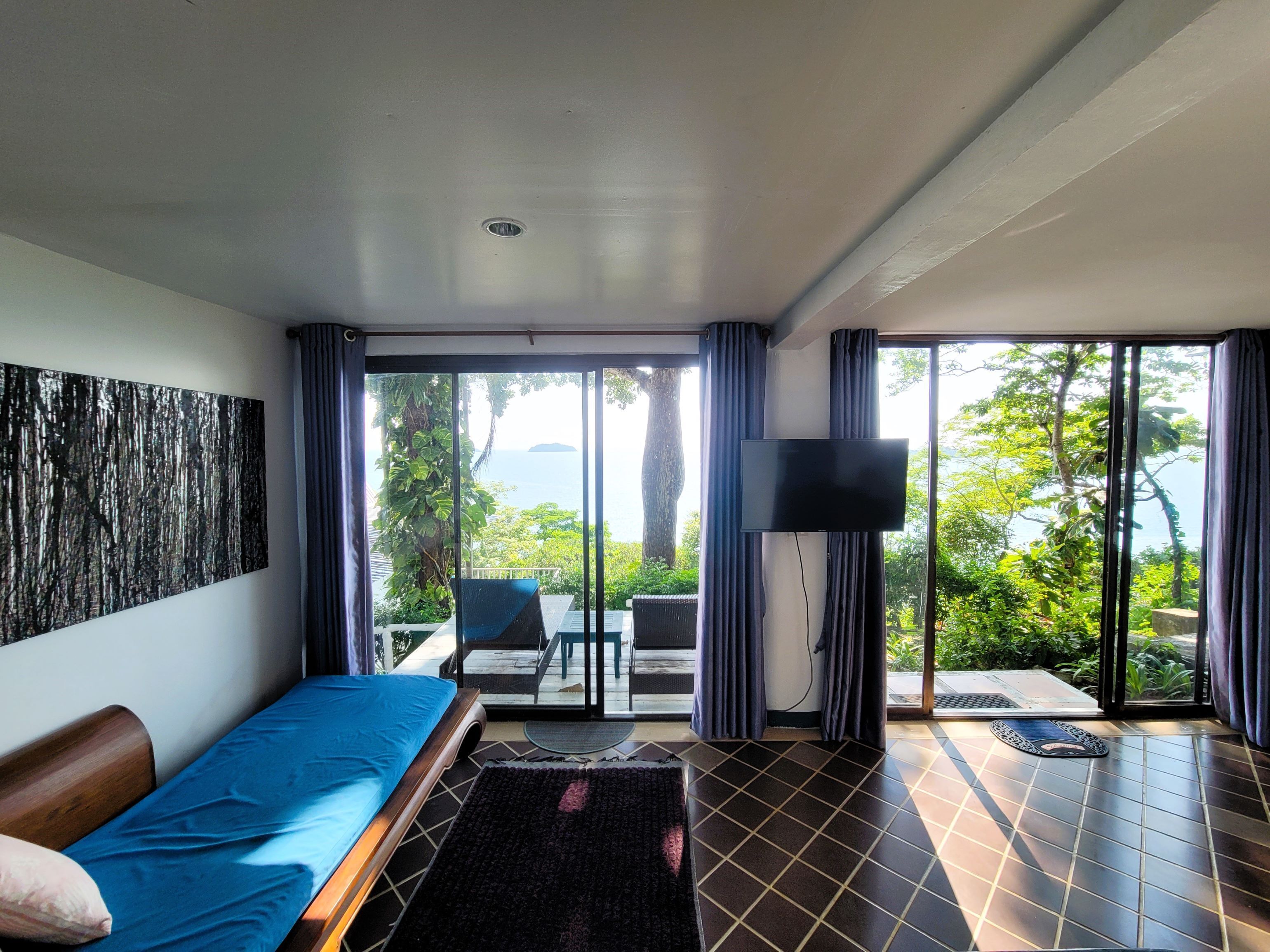 A modern room with a bed, a TV, and a balcony with a view of greenery and the sea.