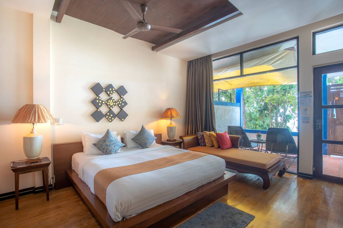 A hotel room with a large bed, two bedside tables, a ceiling fan, a window with curtains, and a balcony with a table and chairs.
