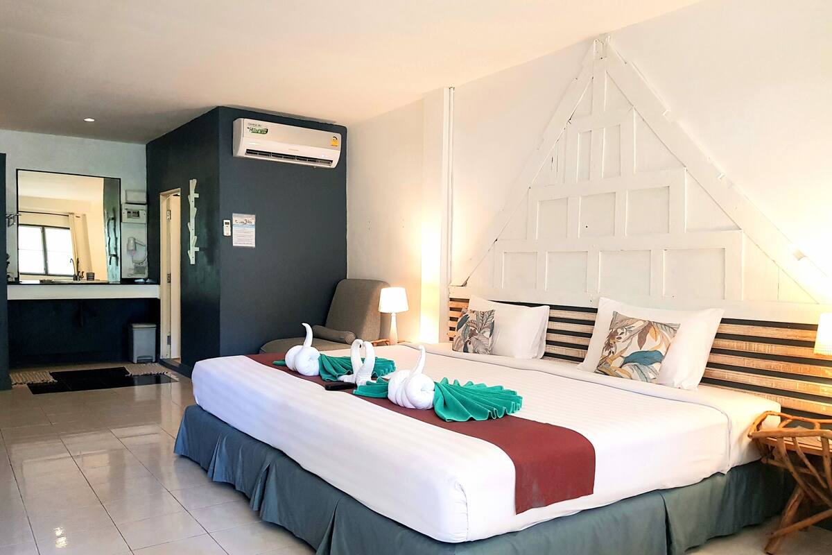 A hotel room with a bed, a desk, a chair, a lamp, and an air conditioning unit.
