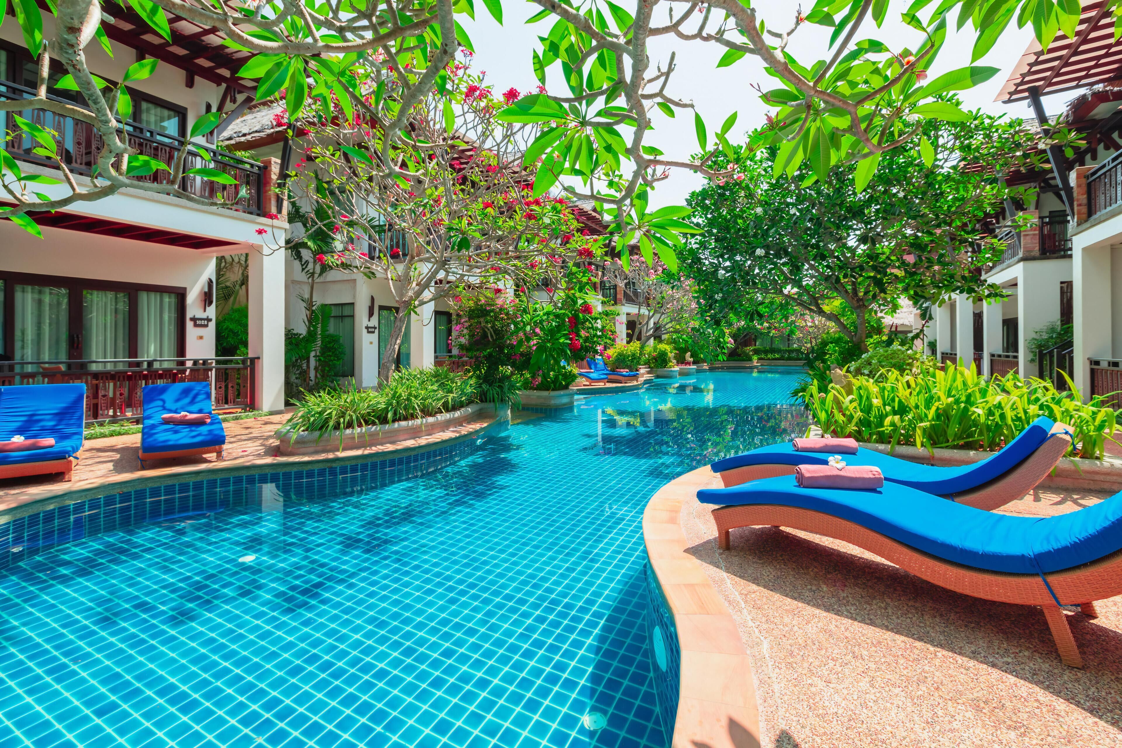 A swimming pool with lounge chairs, surrounded by greenery and a building with balconies.