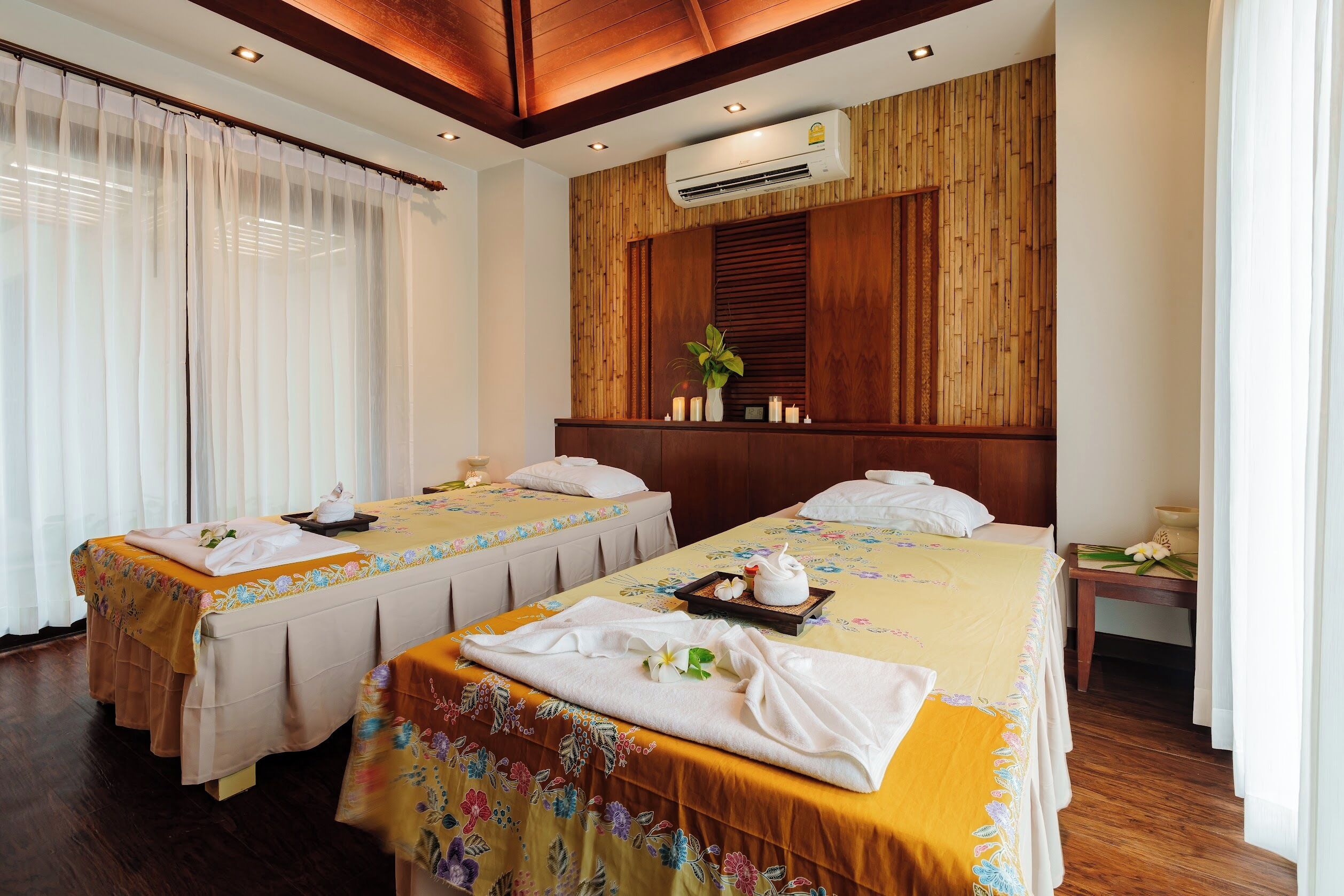 Habitación con dos camas, cada una con cubrecama floral y una toalla. Se aprecia panelado de madera, un equipo de aire acondicionado y una pequeña mesa con una planta y velas.