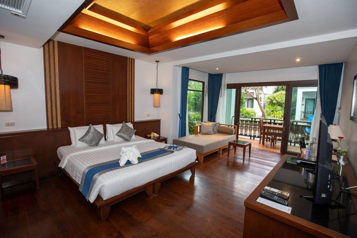 A hotel room with a large bed, a sofa, a desk with a computer, and a balcony with a view of greenery.