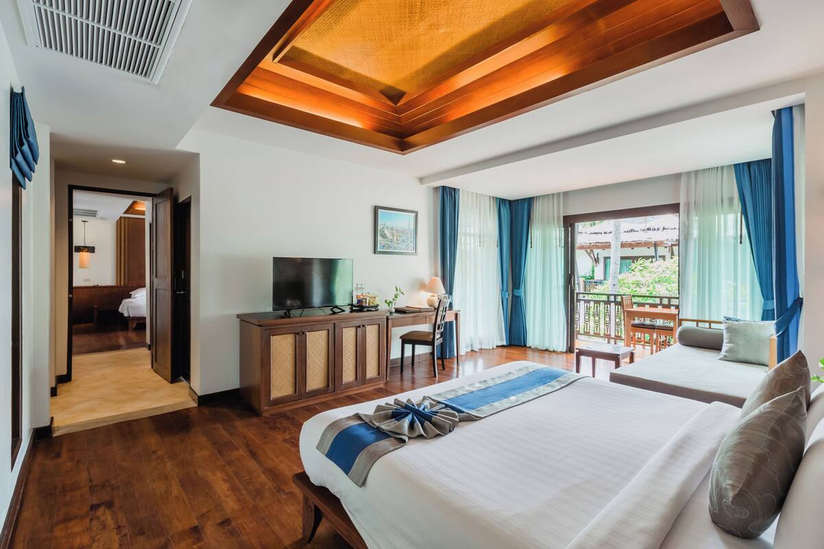 A hotel room with a large bed, a desk with a TV, a balcony with blue curtains, and a view of greenery outside.