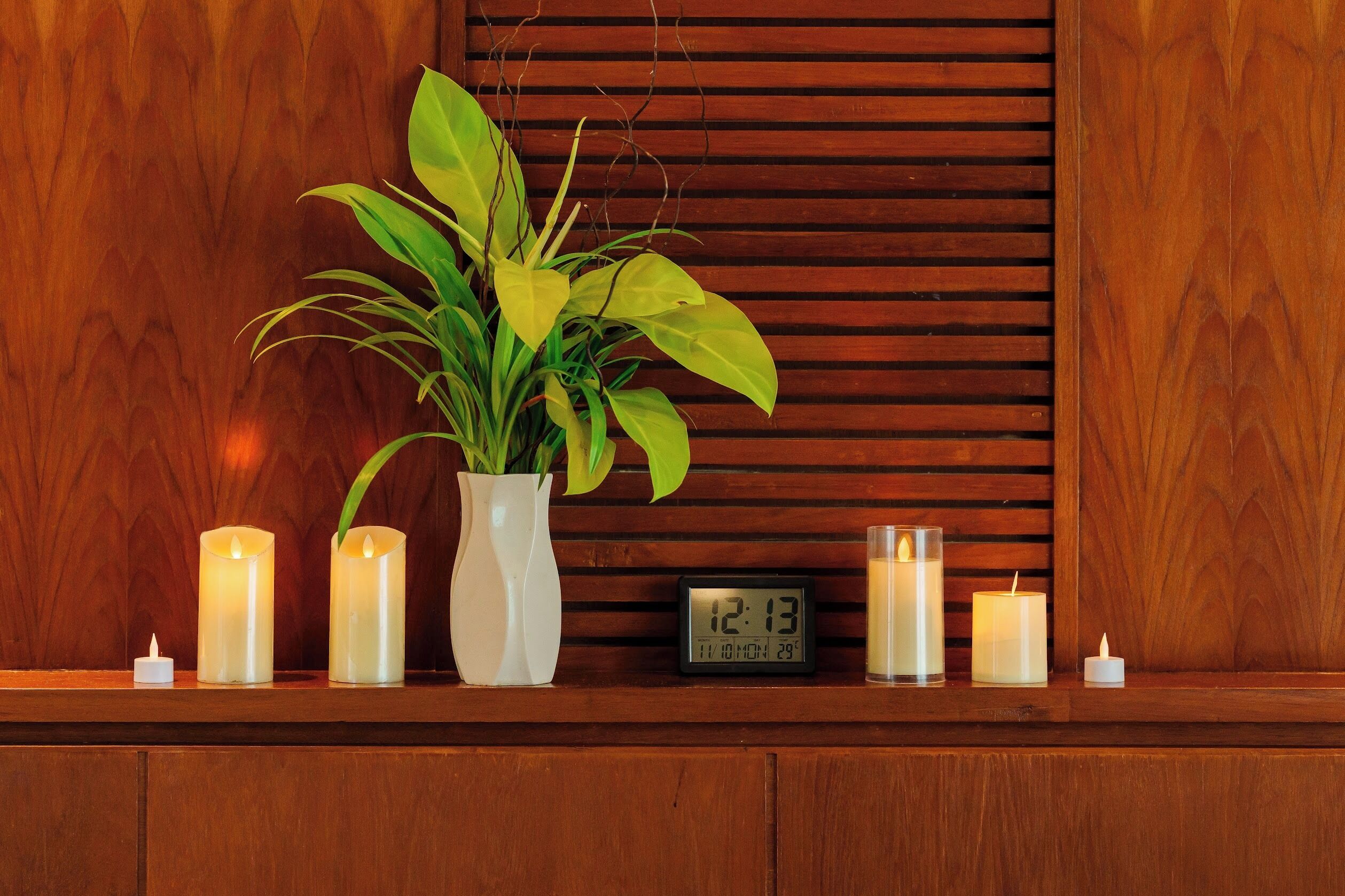 A wooden shelf with candles, a digital clock, and a potted plant.
