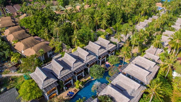 Vista aérea de un complejo turístico con bungalows sobre el agua, una piscina y vistas al océano y a los acantilados.
