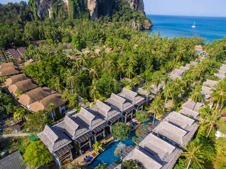 An aerial view of a resort with overwater bungalows, a swimming pool, and a view of the ocean and cliffs.