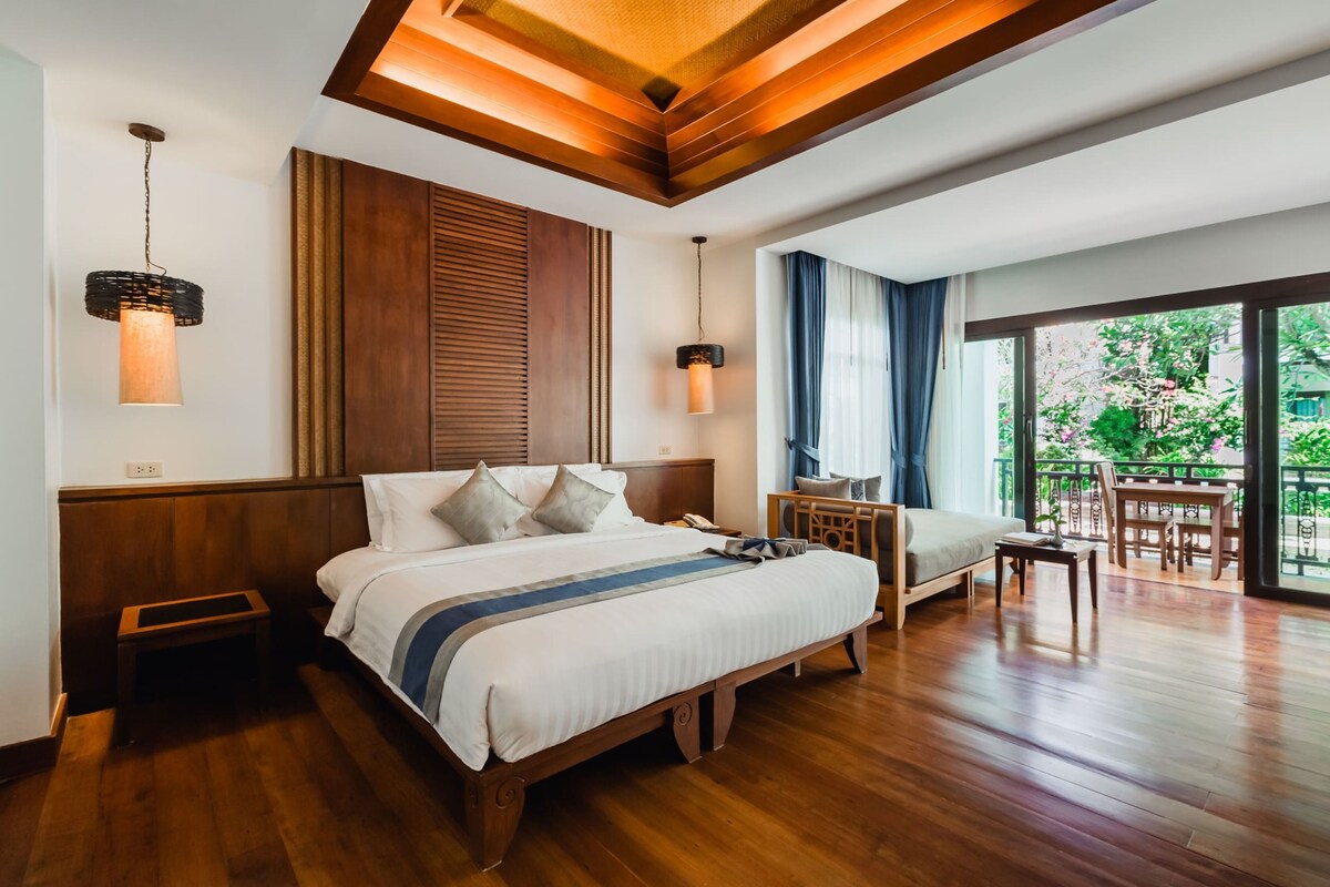 A hotel room with a wooden bed, bedside tables, a desk, and a balcony with a view of greenery.