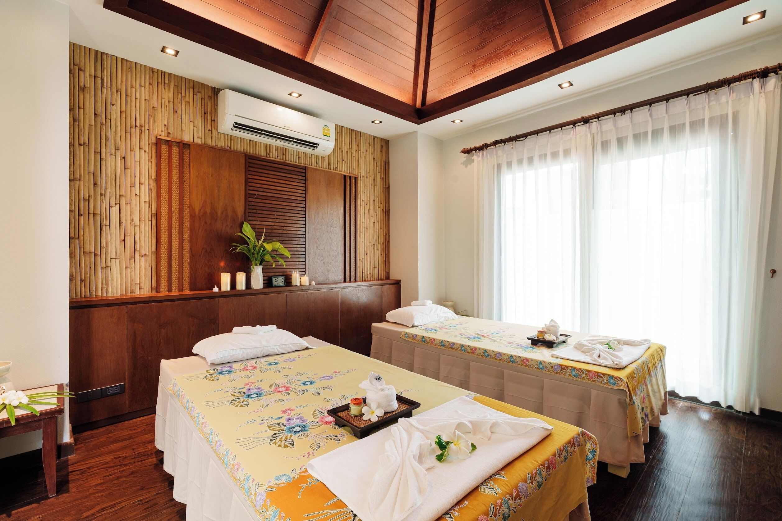 A hotel room with two beds, a wooden wall panel, an air conditioning unit, and a large window with sheer curtains.