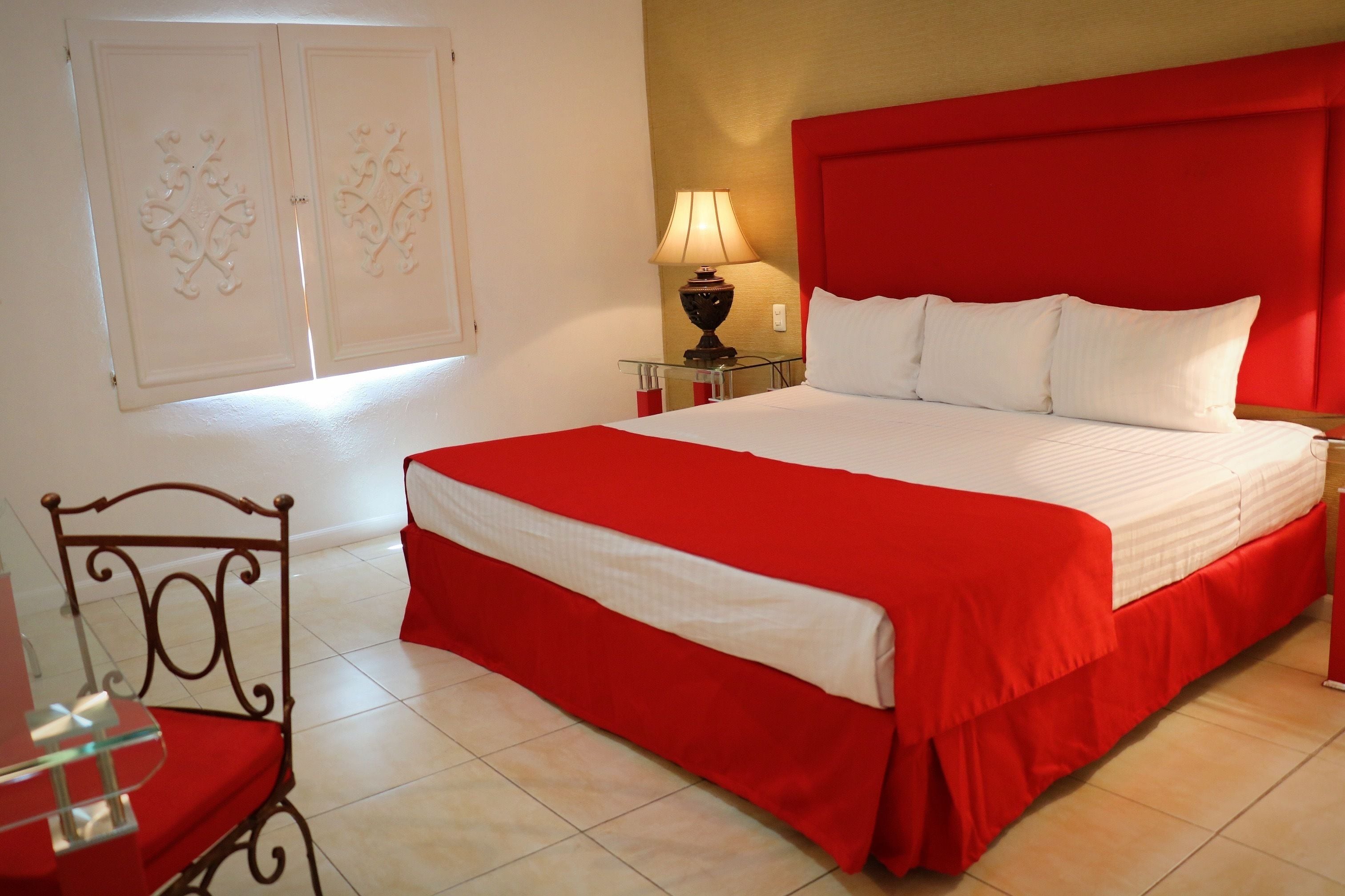 Habitación de hotel con una cama grande, cabecera roja, mesita de noche con lámpara y una silla.