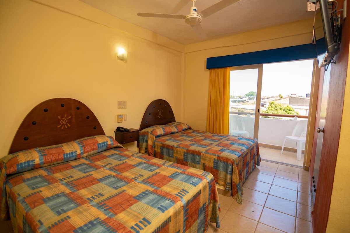 Une chambre d’hôtel avec deux lits, un ventilateur de plafond, une fenêtre avec des rideaux jaunes et une vue sur l’extérieur.