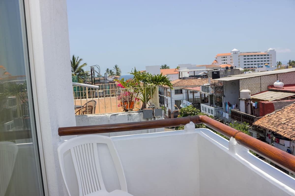 Un balcon avec une chaise en plastique blanche, une balustrade en bois et une vue sur un quartier résidentiel avec des immeubles et des palmiers.