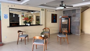 Un área de recepción con un mostrador, tres sillas de madera con cojines grises, un espejo grande, ventiladores de techo y un panel decorativo en la pared.