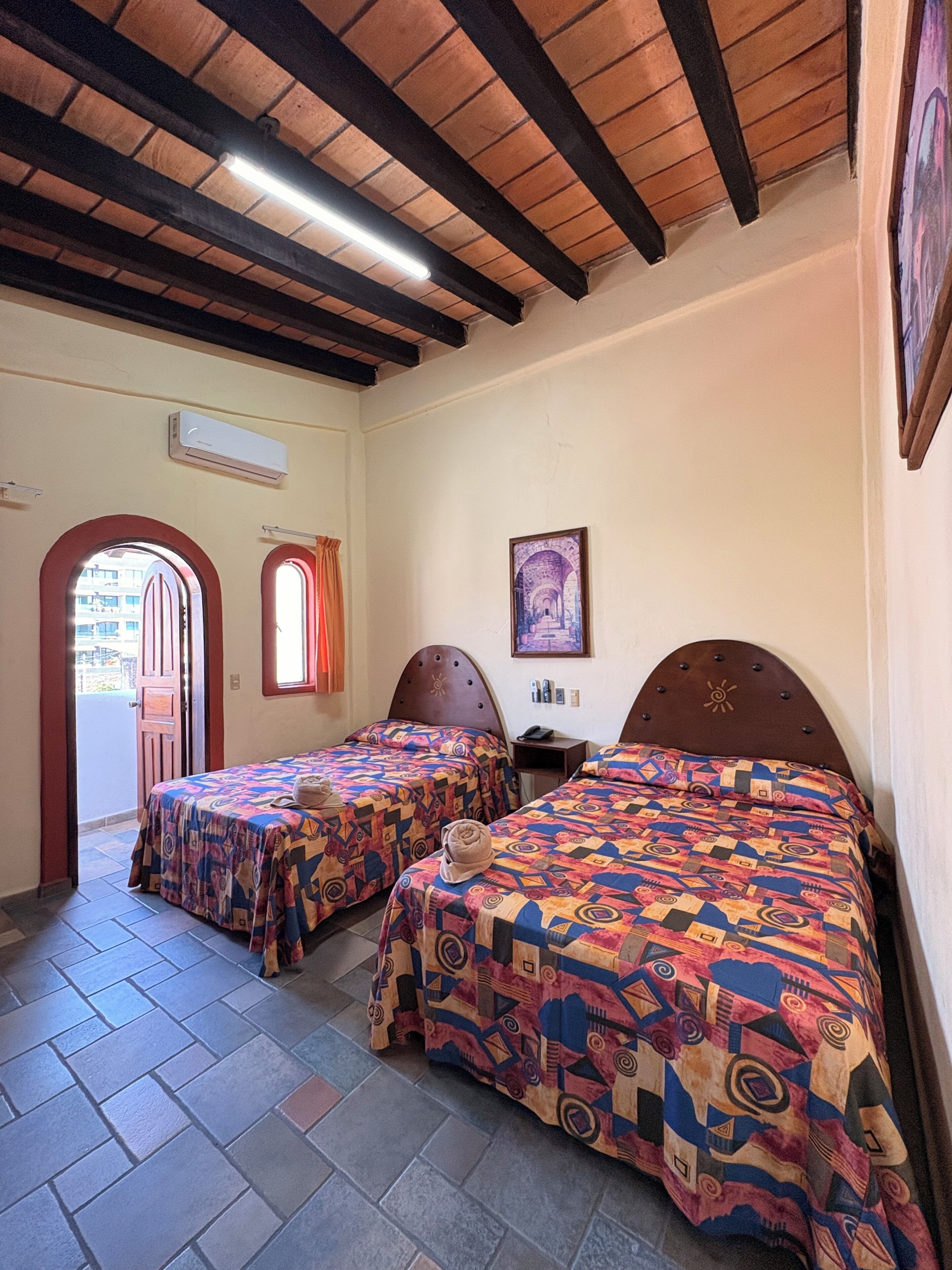 Una habitación con dos camas, techo de madera, unidad de aire acondicionado, un cuadro en la pared y una ventana con cortinas.
