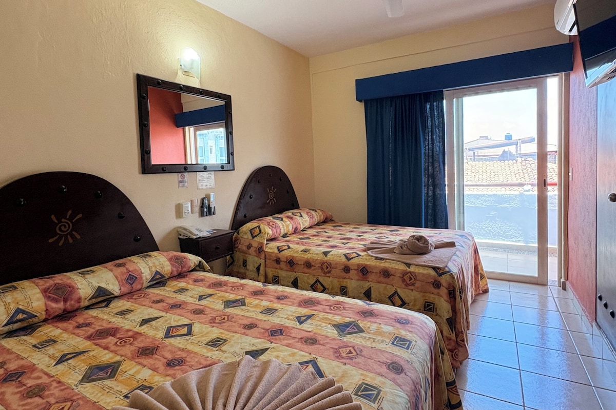 Habitación de hotel con dos camas, ventilador de techo, ventana con cortinas y vistas al exterior.
