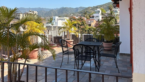 Un balcón con vistas a un paisaje urbano, que incluye una mesa, sillas, plantas en macetas y una barandilla.