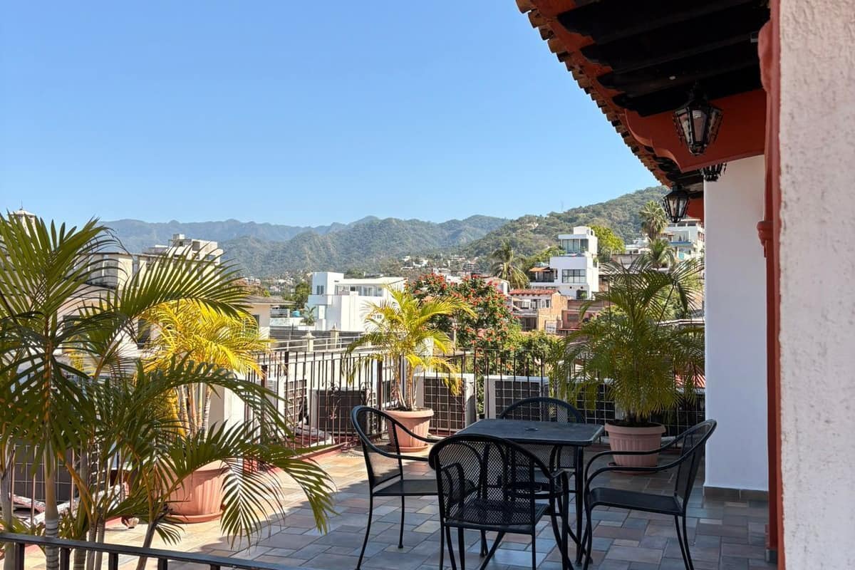 Varanda com vista para a cidade, com mesa, cadeiras, plantas em vasos e grade.
