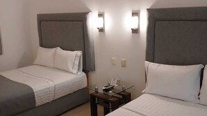 Habitación de hotel con dos camas, cada una con cabecero gris y ropa de cama blanca. Hay una mesita con una lámpara y un control remoto.