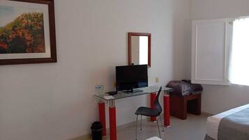 A hotel room with a glass desk, a television, a chair, a bed, a window, and a painting on the wall.