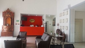 Una zona de recepción con un mostrador rojo, dos sofás de mimbre, un aparador de madera y suelo de baldosas.