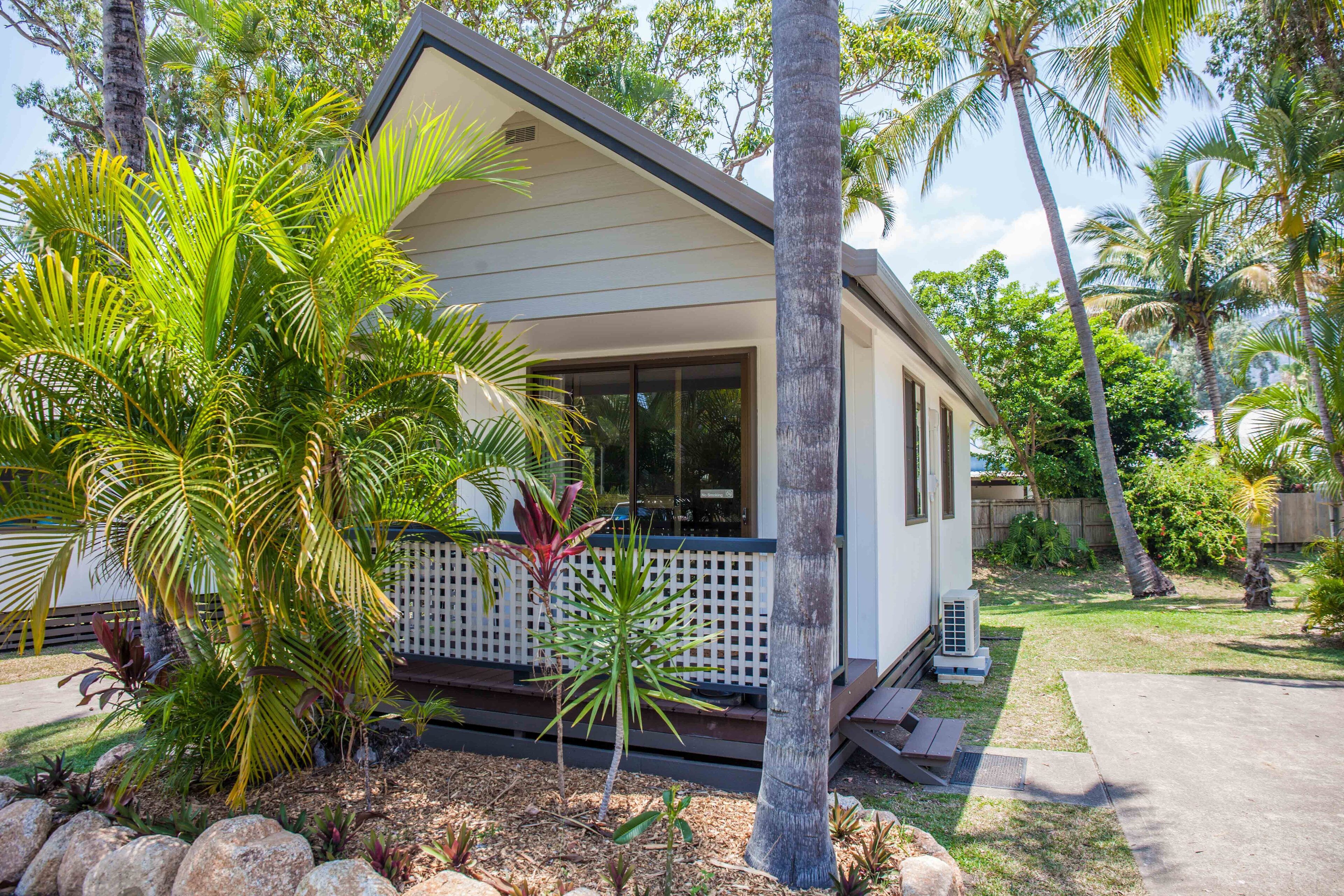 Une maison de plain-pied dotée d’un porche abrité, entourée d’une végétation tropicale et agrémentée d’une terrasse en bois.