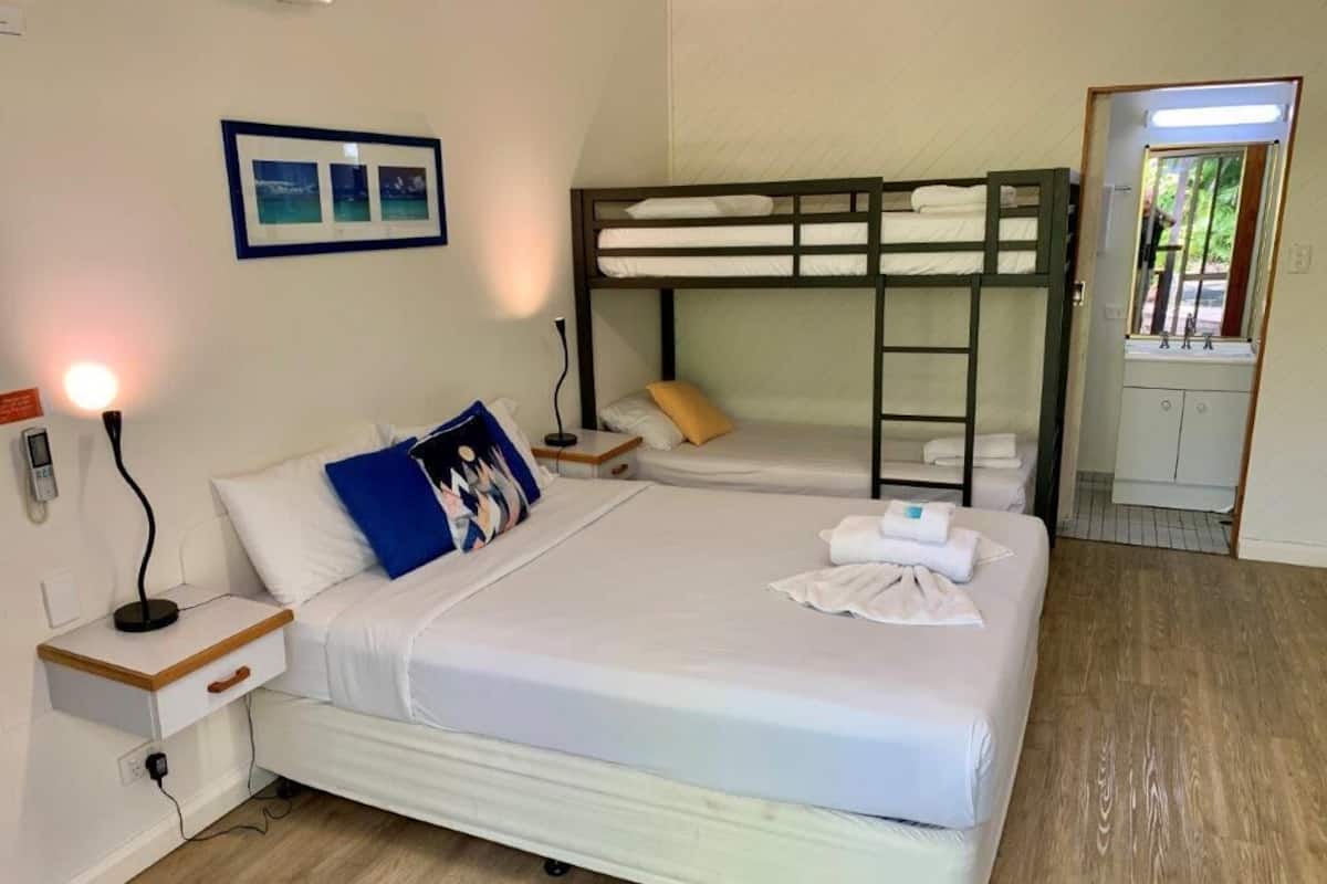 Une chambre avec un lit superposé, une table de chevet avec une lampe, un tableau encadré au mur et une salle de bain avec un miroir et un lavabo.