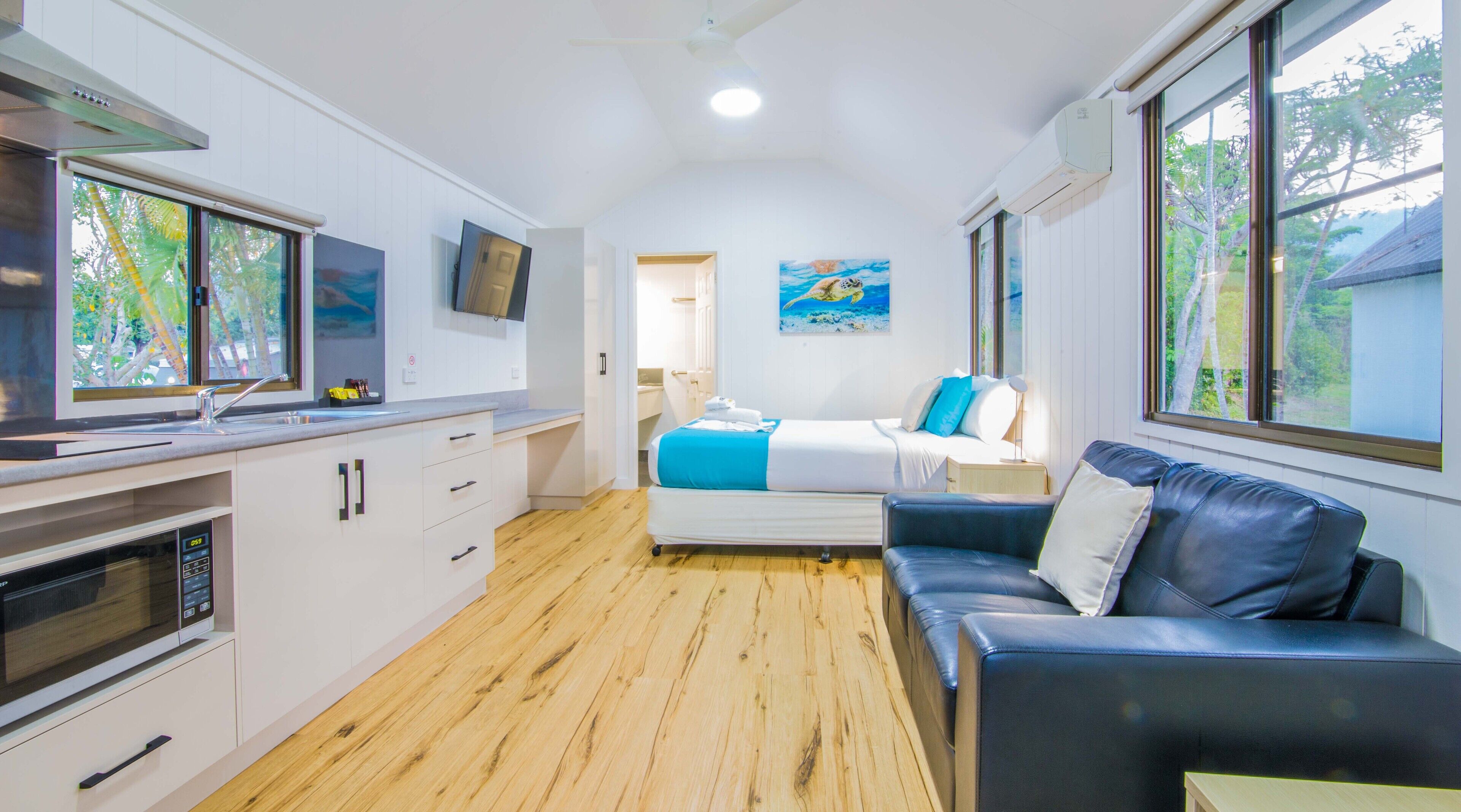 A compact hotel room with a kitchenette, a bed with blue bedding, a leather sofa, and large windows offering a view of trees.